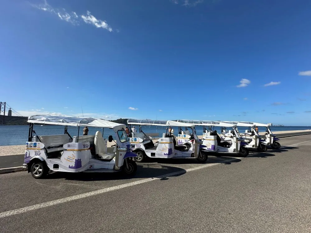 Fleet of Just Tour It electric Tuk Tuks lined up along Lisbon’s Tagus River with the 25 de Abril Bridge in the background.