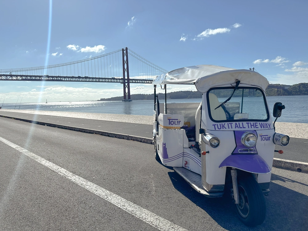 Electric Tuk Tuk from Just Tour It parked by the Tagus River with the 25 de Abril Bridge in the background on a sunny day in Lisbon.