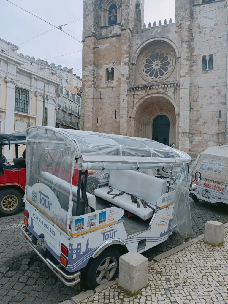 Just Tour It electric Tuk Tuk parked in front of the historic Lisbon Cathedral (Sé de Lisboa) on a cobblestone street.