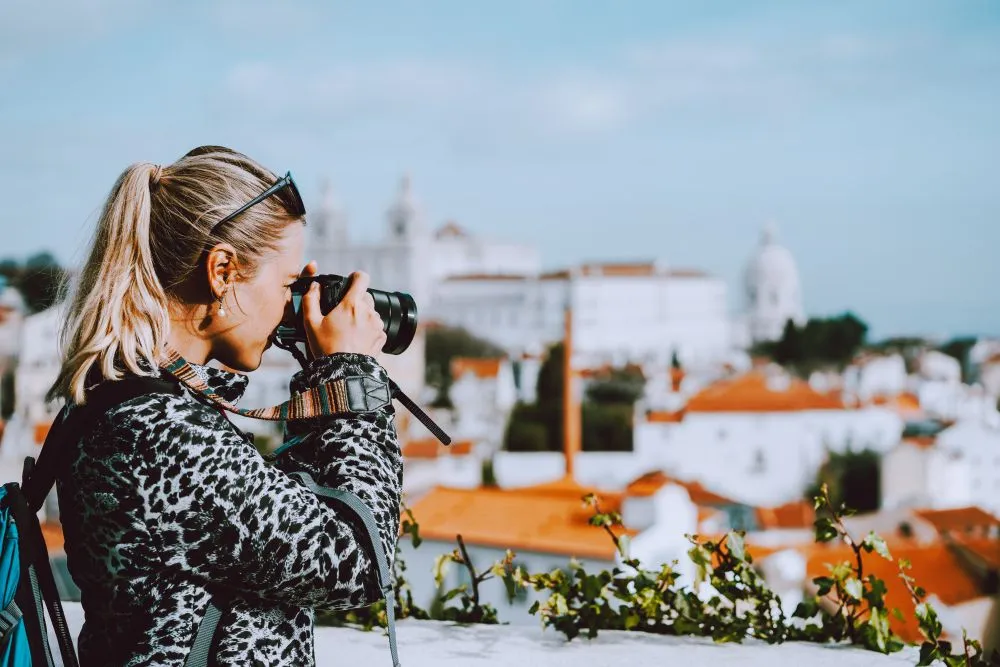 Female traveler taking a photo of Lisbon’s red rooftops and historic skyline from a scenic city viewpoint.